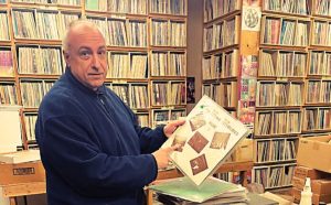 Gregg Kostelich of Get Hip Records standing in front of shelves full of records while holding one up and pointing at it.