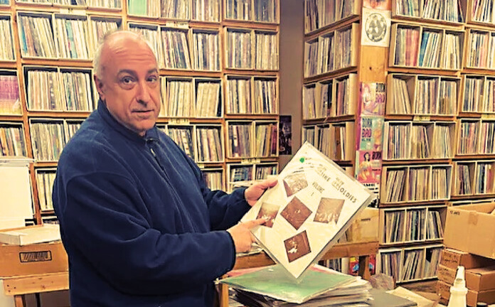 Gregg Kostelich of Get Hip Records standing in front of shelves full of records while holding one up and pointing at it.